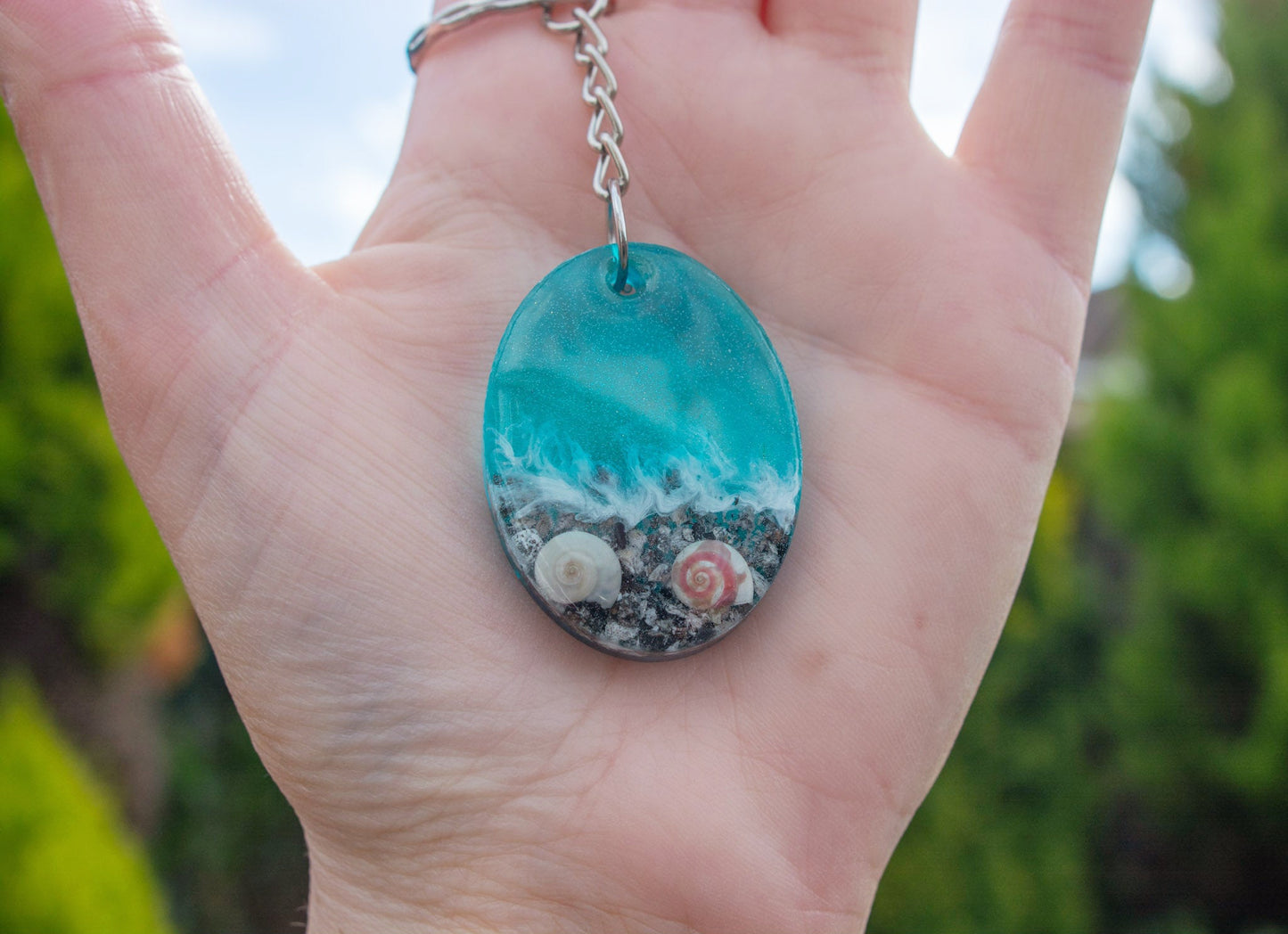 A hand holding an Oval shaped keepsake memorial keyring outside, in a seashore theme, containing sand, cremation ashes and shells