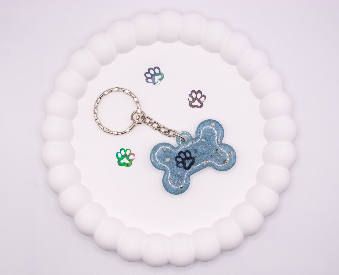 An aerial, overhead view of a light blue bone shaped keepsake memorial keyring, containing pet cremation ashes, set on a silver keychain