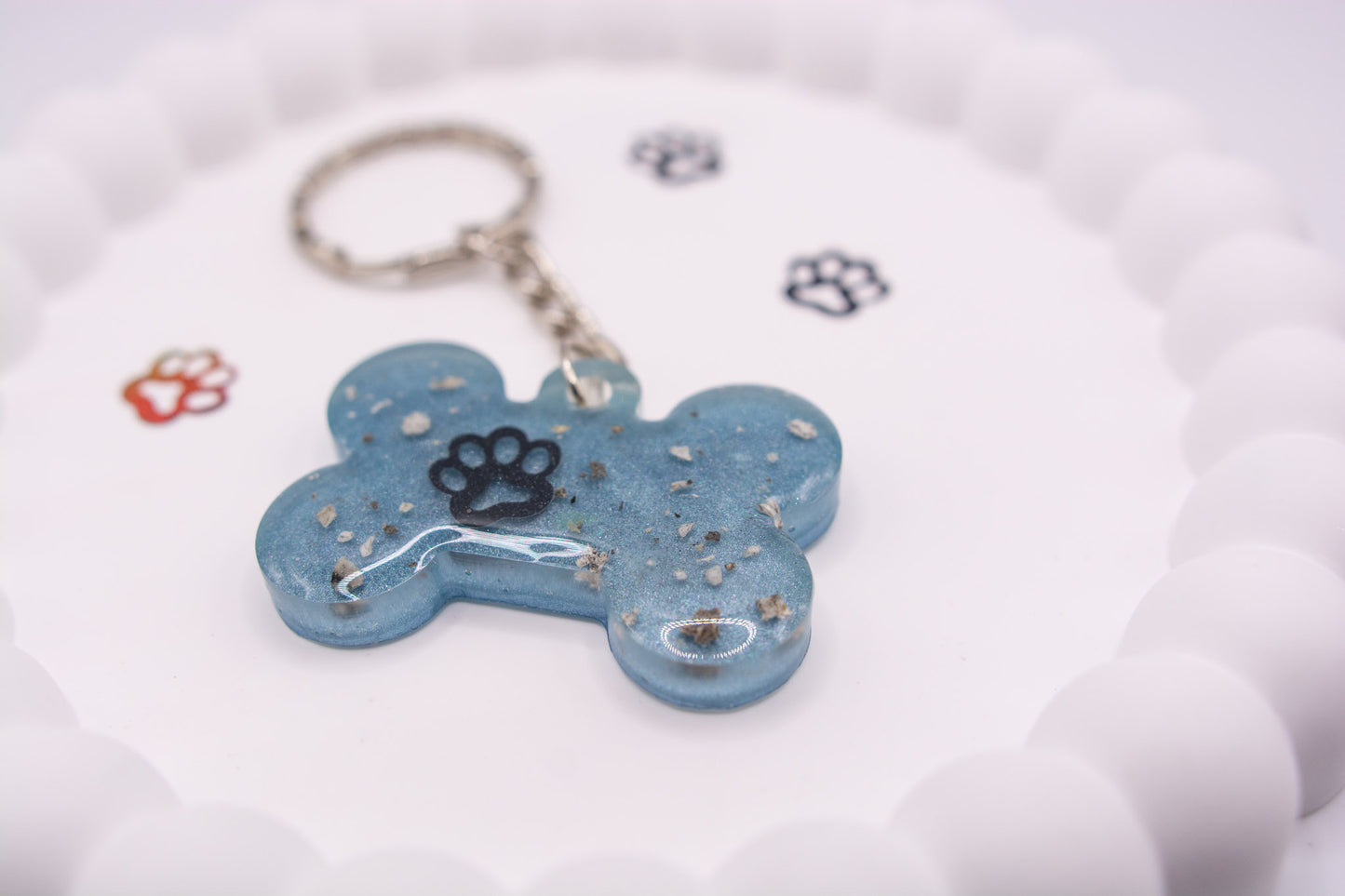 A close up angled view of a light blue bone shaped keyring, containing a paw print decal and dog cremation ashes set on a silver keychain