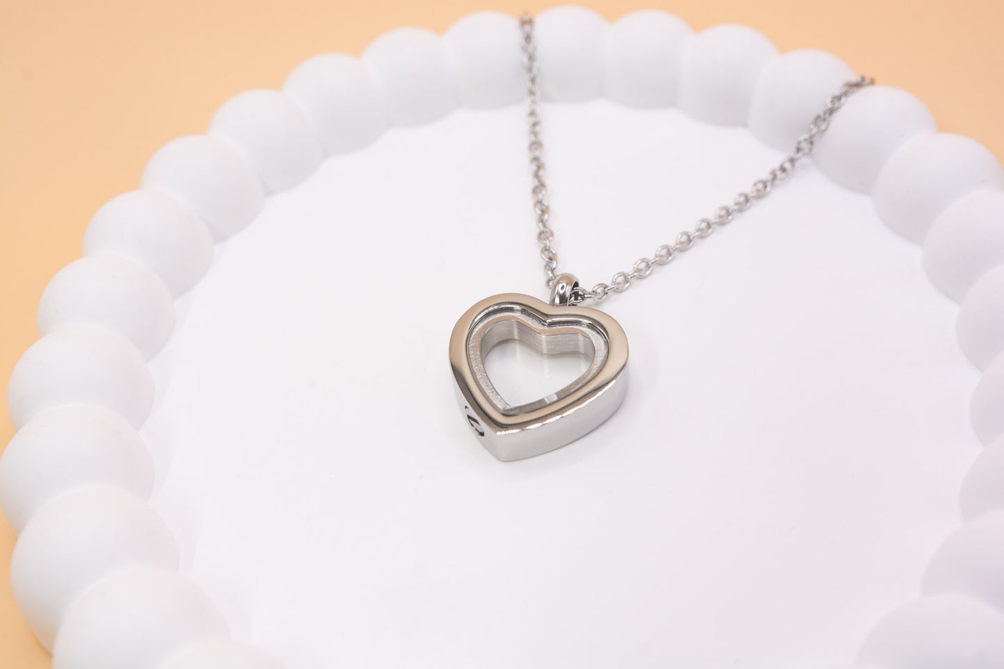 An angled close up view of a stainless steel heart pendant, which can be self-filled with cremation ashes or sand from a favourite beach, it has a clear central window so you can see the contents