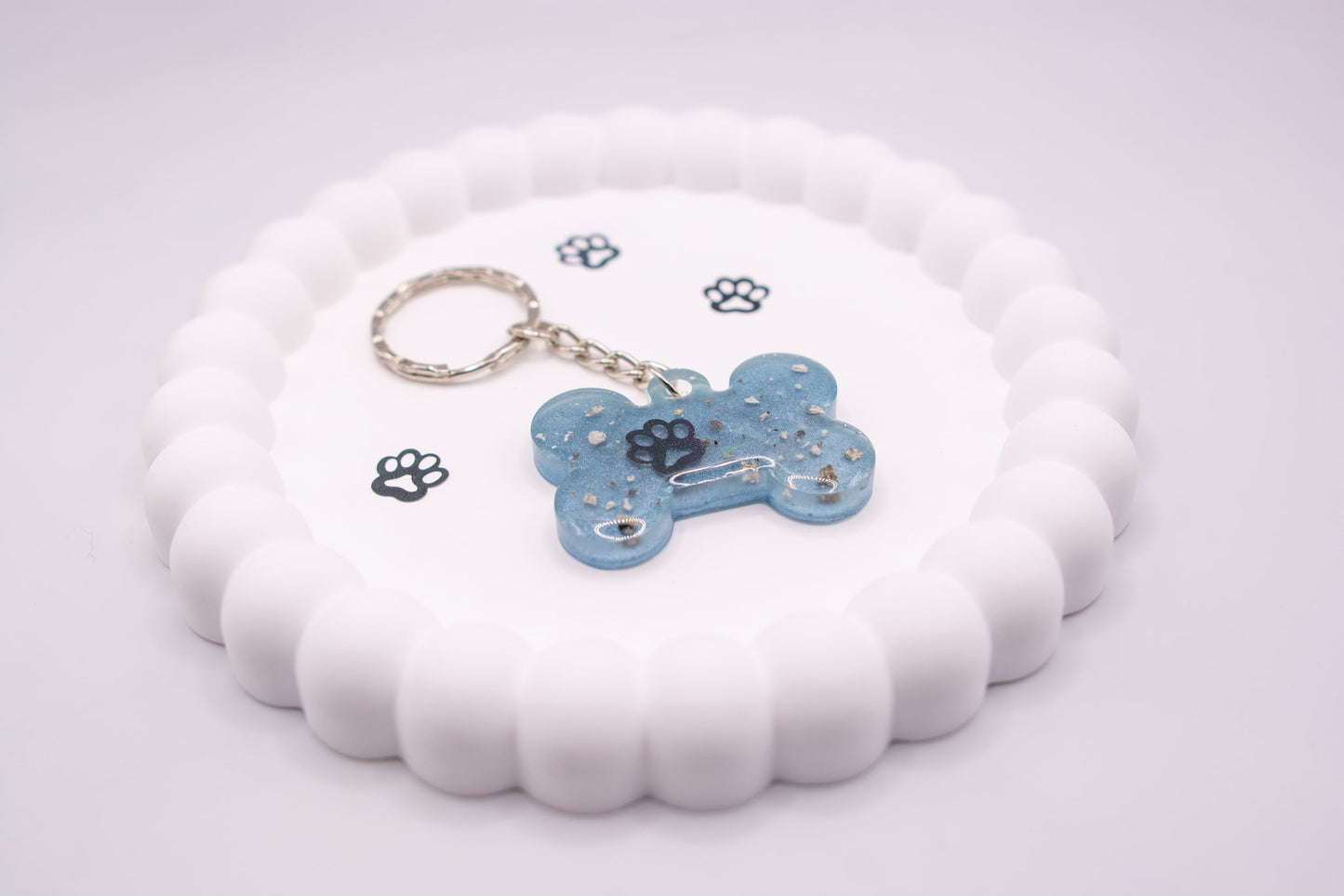 Angled view of a light blue bone shaped keepsake memorial keyring, containing a paw decoration and pet ashes, set on a silver keychain and presented on a display tray