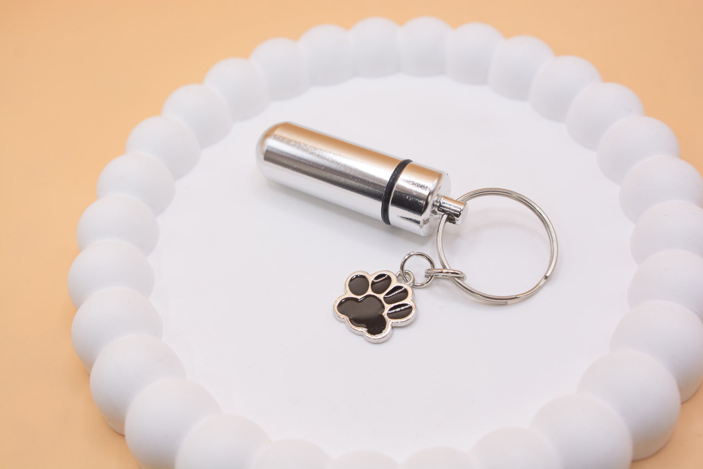 An angled close up view of a stainless steel silver keyring, with a black paw print charm. The keyring can be filled with a lost loved pet such as a cat or dog's cremation ashes. Displayed on a white tray