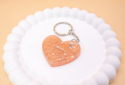 A Rose Gold heart shaped keyring containing cremation ashes with the word Mum on it, presented on a display tray, angled close view