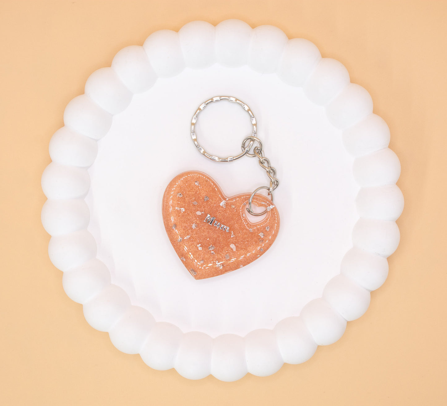 A Rose Gold heart shaped keyring containing cremation ashes with the word "Mum" on it, presented on a display tray, overhead view
