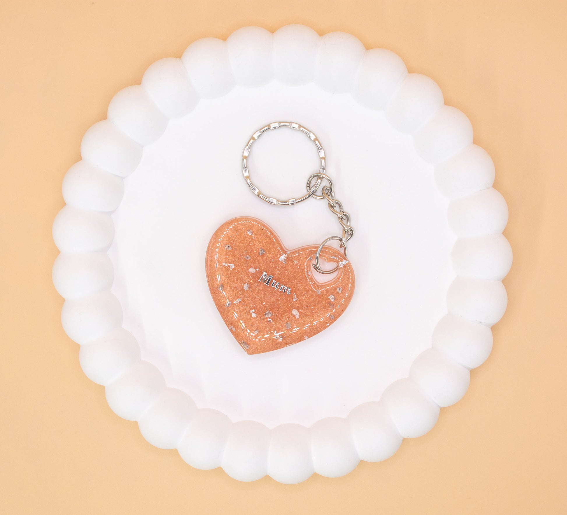 A Rose Gold heart shaped keyring containing cremation ashes with the word "Mum" on it, presented on a display tray, overhead view