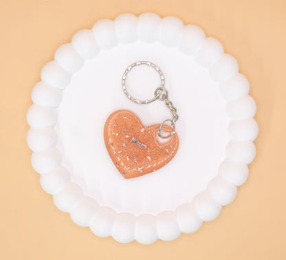 A Rose Gold heart shaped keyring containing cremation ashes with the word "Mum" on it, presented on a display tray, overhead view