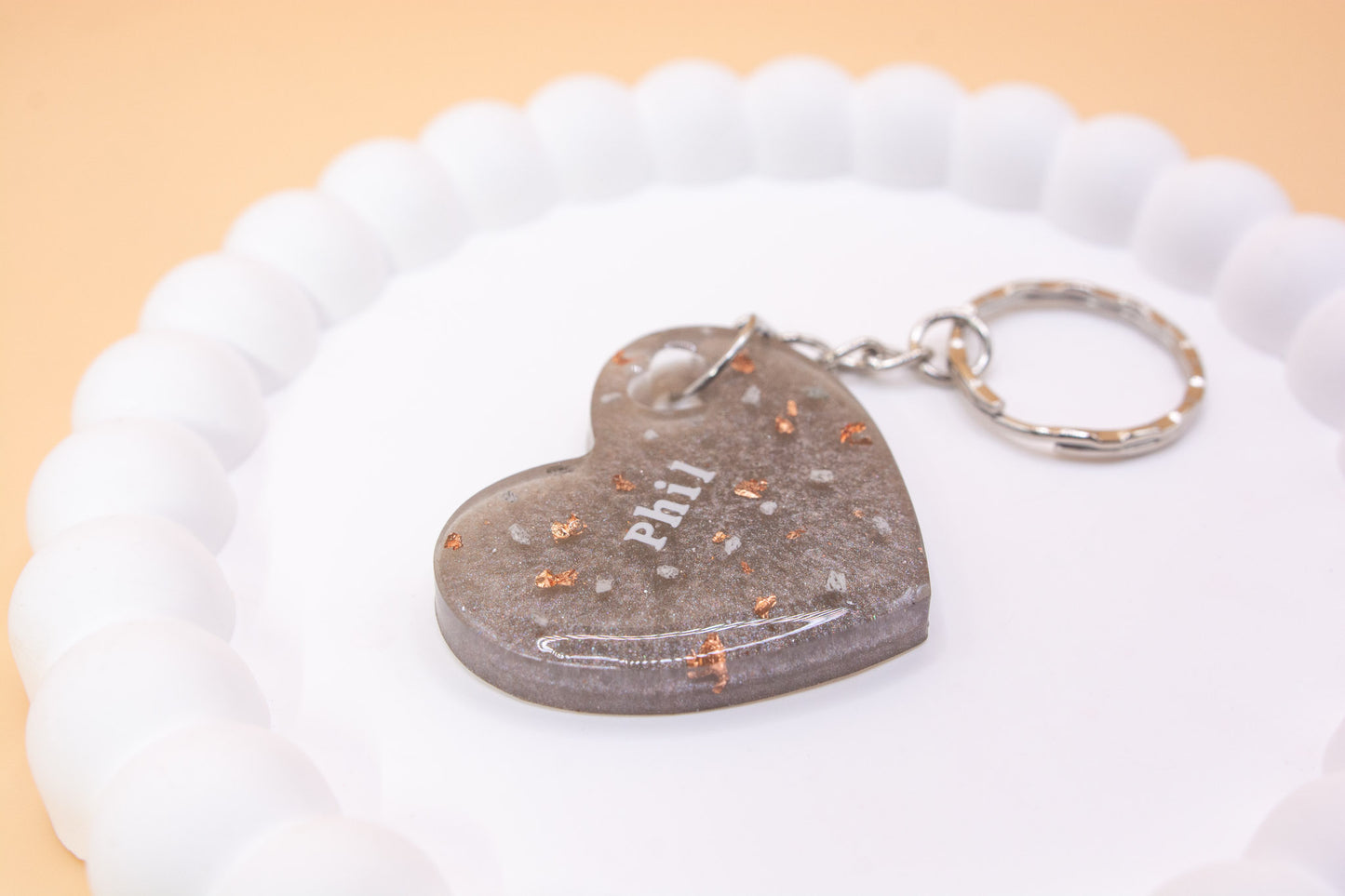 A Metallic Silver Grey heart shaped keyring containing cremation ashes with the name "Phil" on it, presented on a display tray, close up view