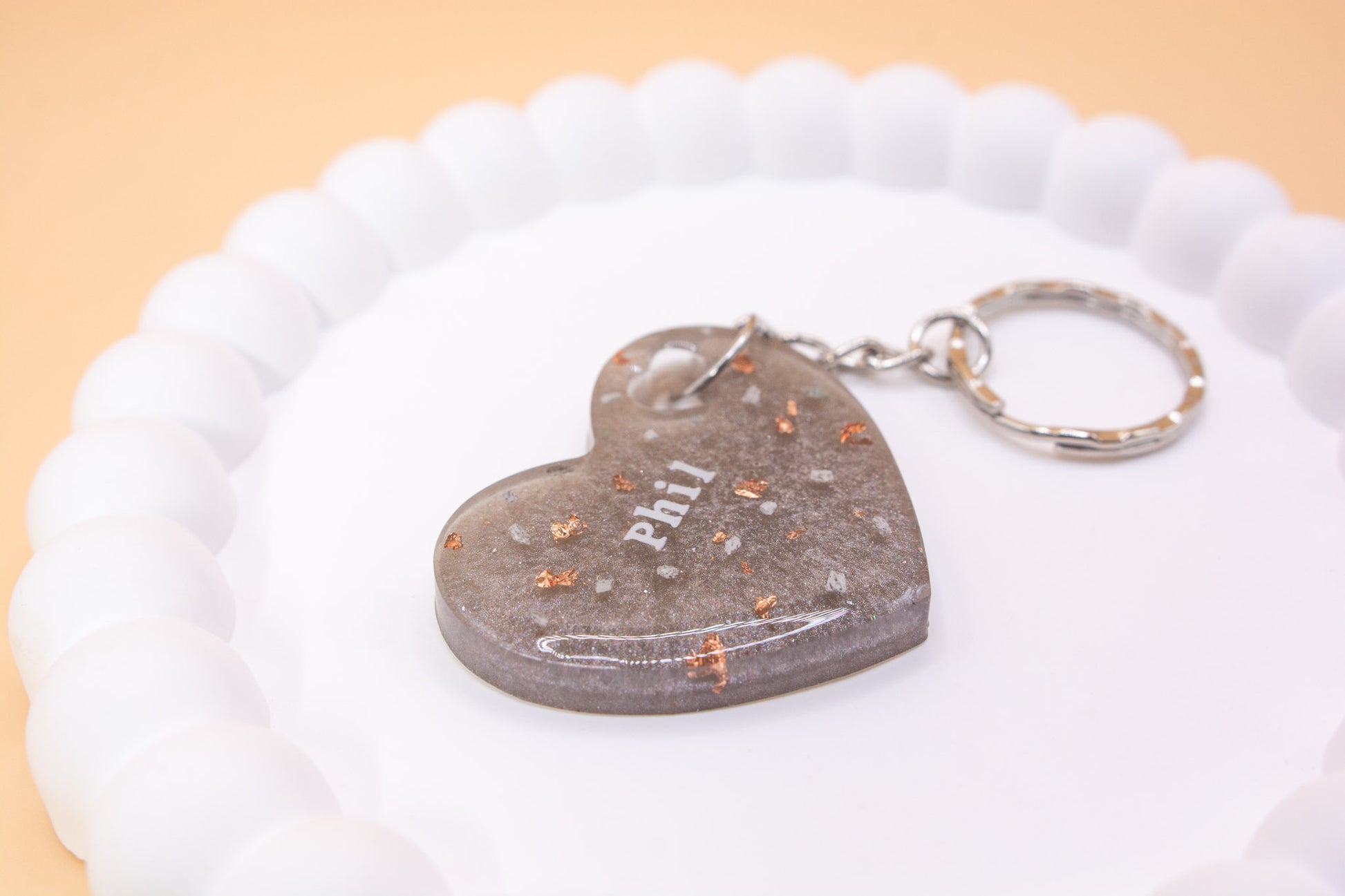A Metallic Silver Grey heart shaped keyring containing cremation ashes with the name "Phil" on it, presented on a display tray, close up view