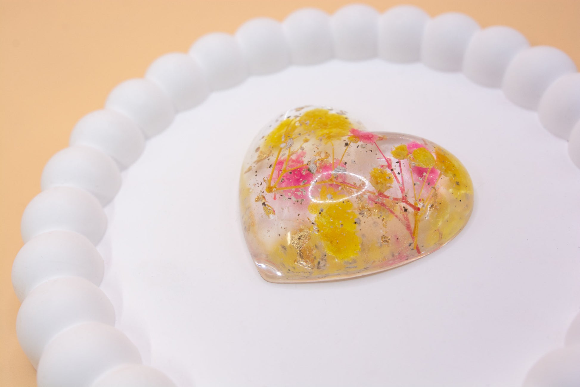 A close up view of a heart shaped ornament containing cremation ashes, yellow and pink dried flowers and gold flakes, presented on a display tray