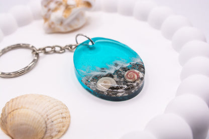A close up view of an Oval keepsake memorial keyring with seashore theme, containing sand, cremation ashes and shells