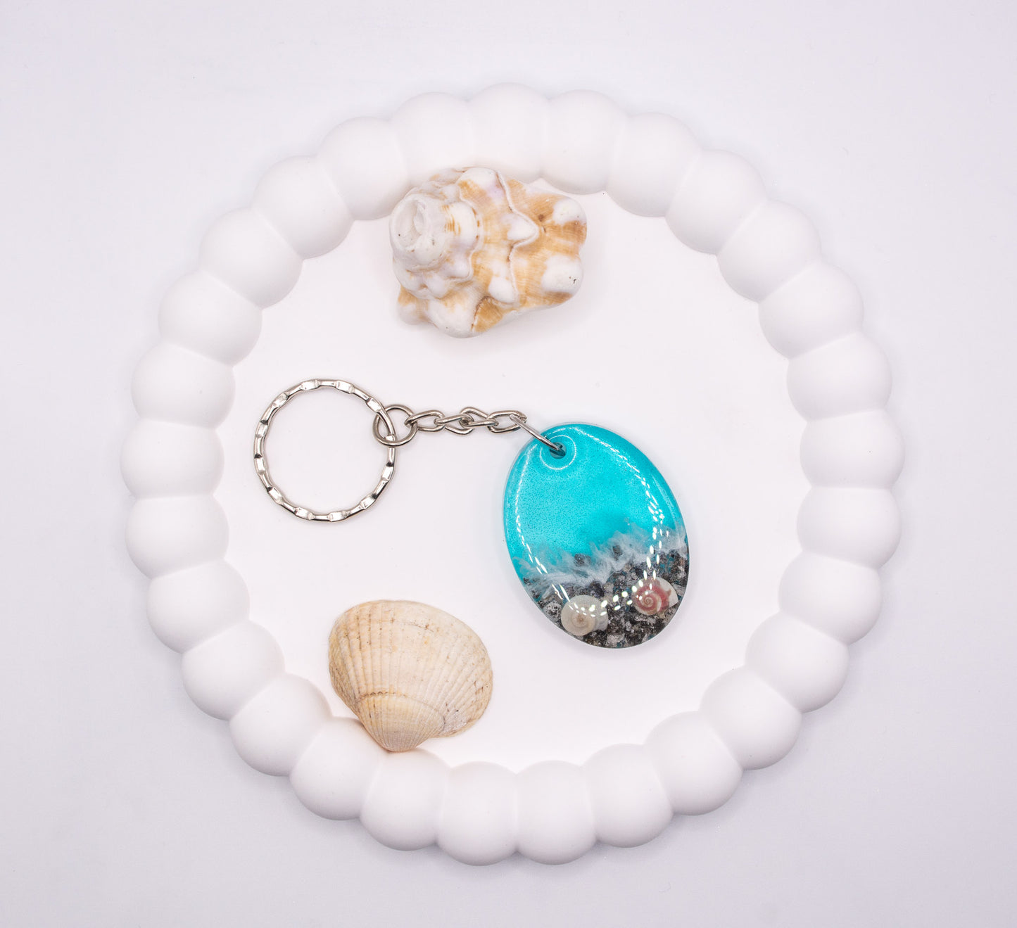 An aerial overhead view of an Oval shaped keepsake memorial keyring with seashore theme, containing sand, cremation ashes and shells, presented next to beach shells, displayed on a jewelry tray