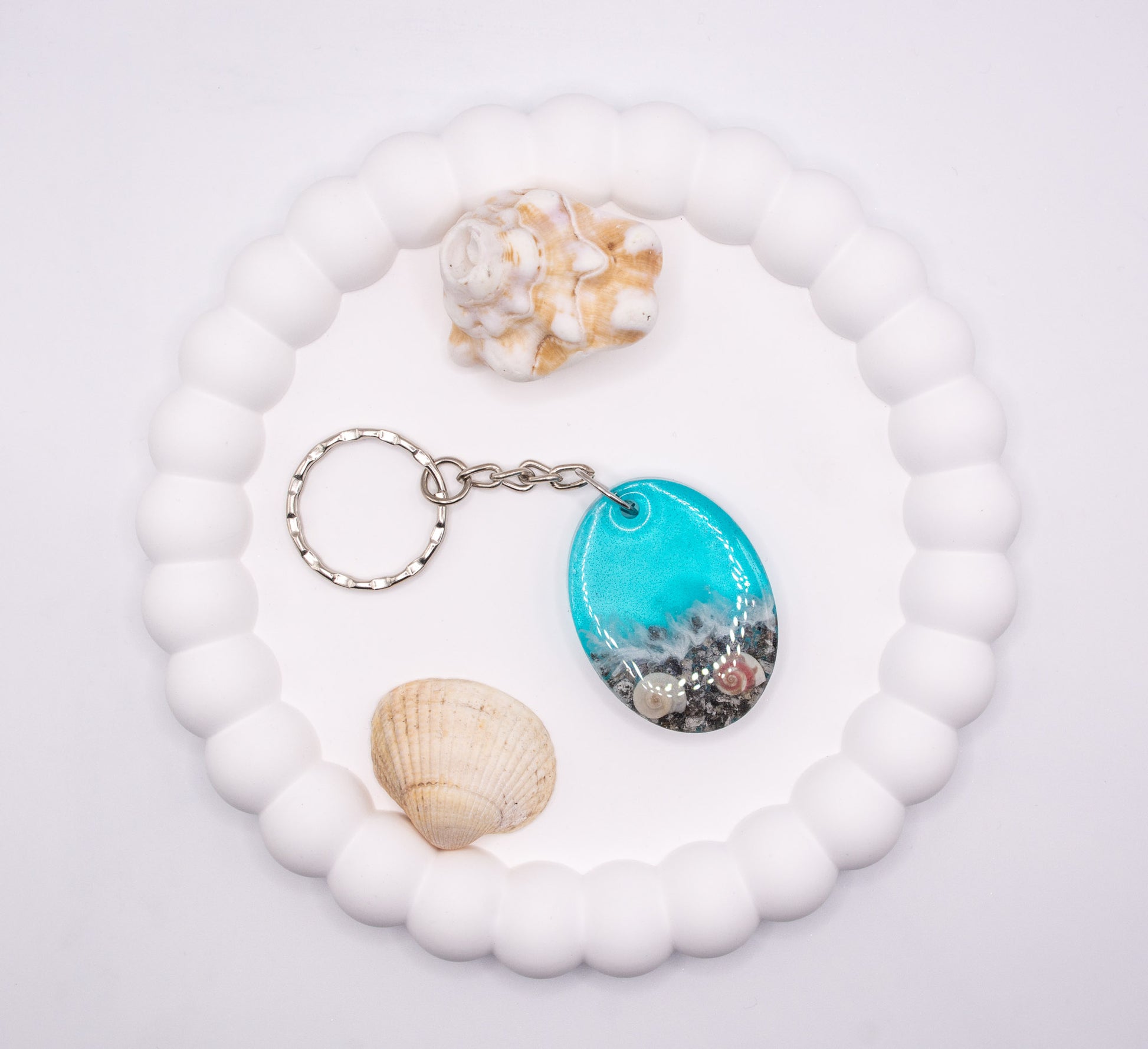 An aerial overhead view of an Oval shaped keepsake memorial keyring with seashore theme, containing sand, cremation ashes and shells, presented next to beach shells, displayed on a jewelry tray