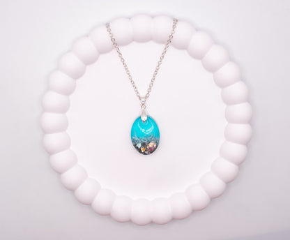 An overhead view of an Oval shaped seashore memorial keepsake pendant, containing cremation ashes and shell pieces, presented on a sterling 925 silver chain
