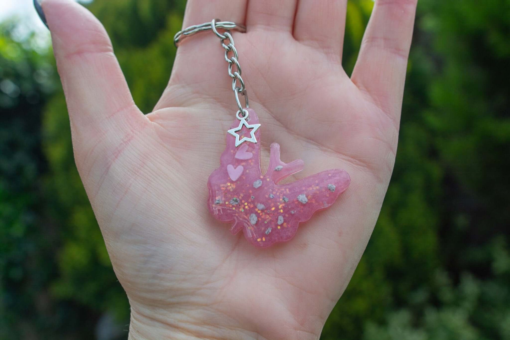 A hand holding a Pink butterfly keepsake memorial keyring outside, which contains cremation ashes and glitter, set on a silver keychain