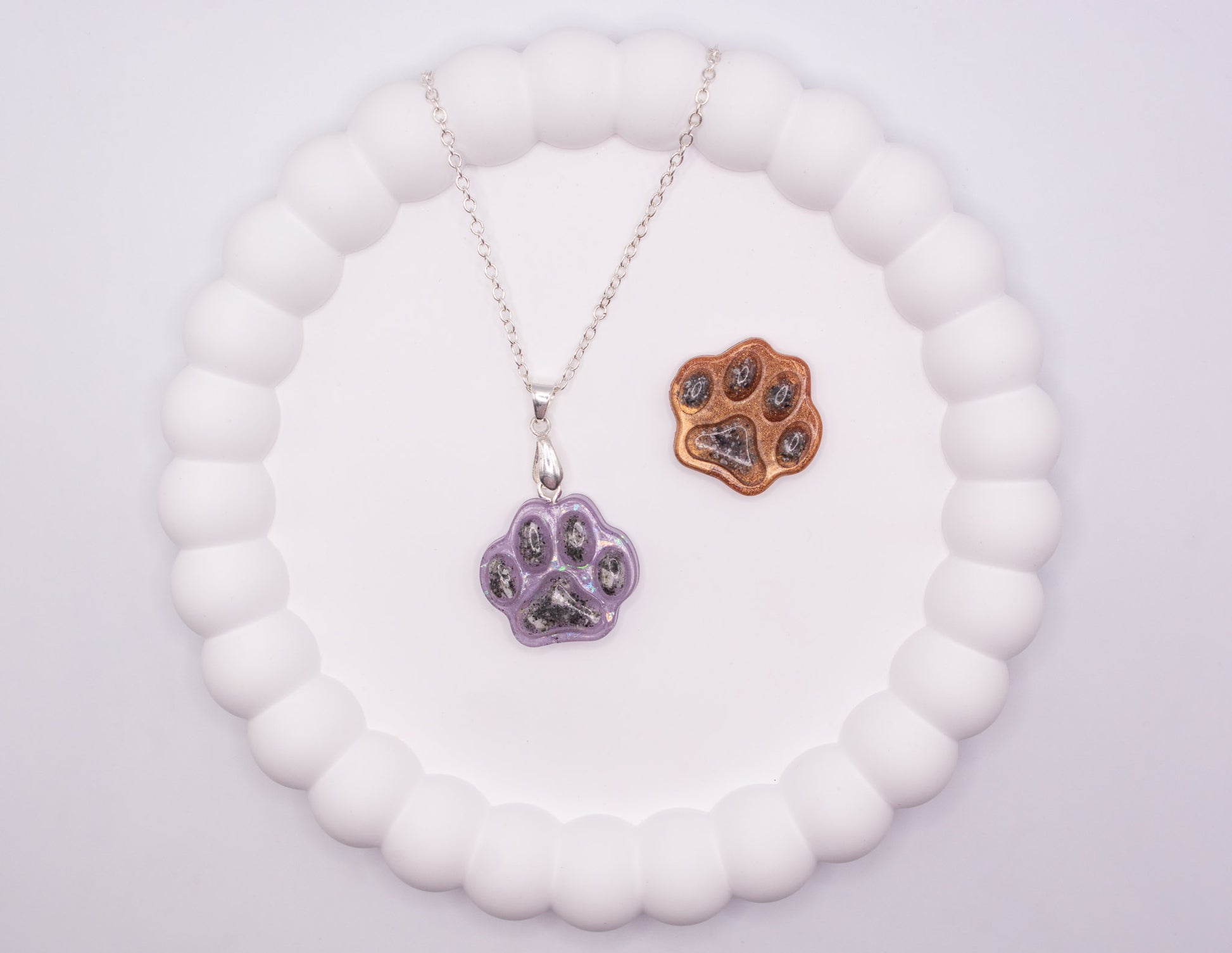An aerial overhead view of 2 Paw prints, one is glitter purple and set on a 925 silver chain, and the other one is metallic brown, both containing pet cremation ashes, and displayed on a jewelry tray