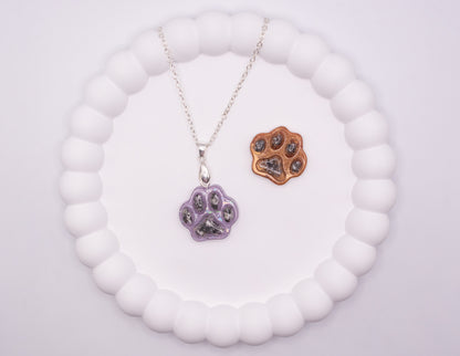 An aerial overhead view of 2 Paw prints, one is glitter purple and set on a 925 silver chain, and the other one is metallic brown, both containing pet cremation ashes, and displayed on a jewelry tray
