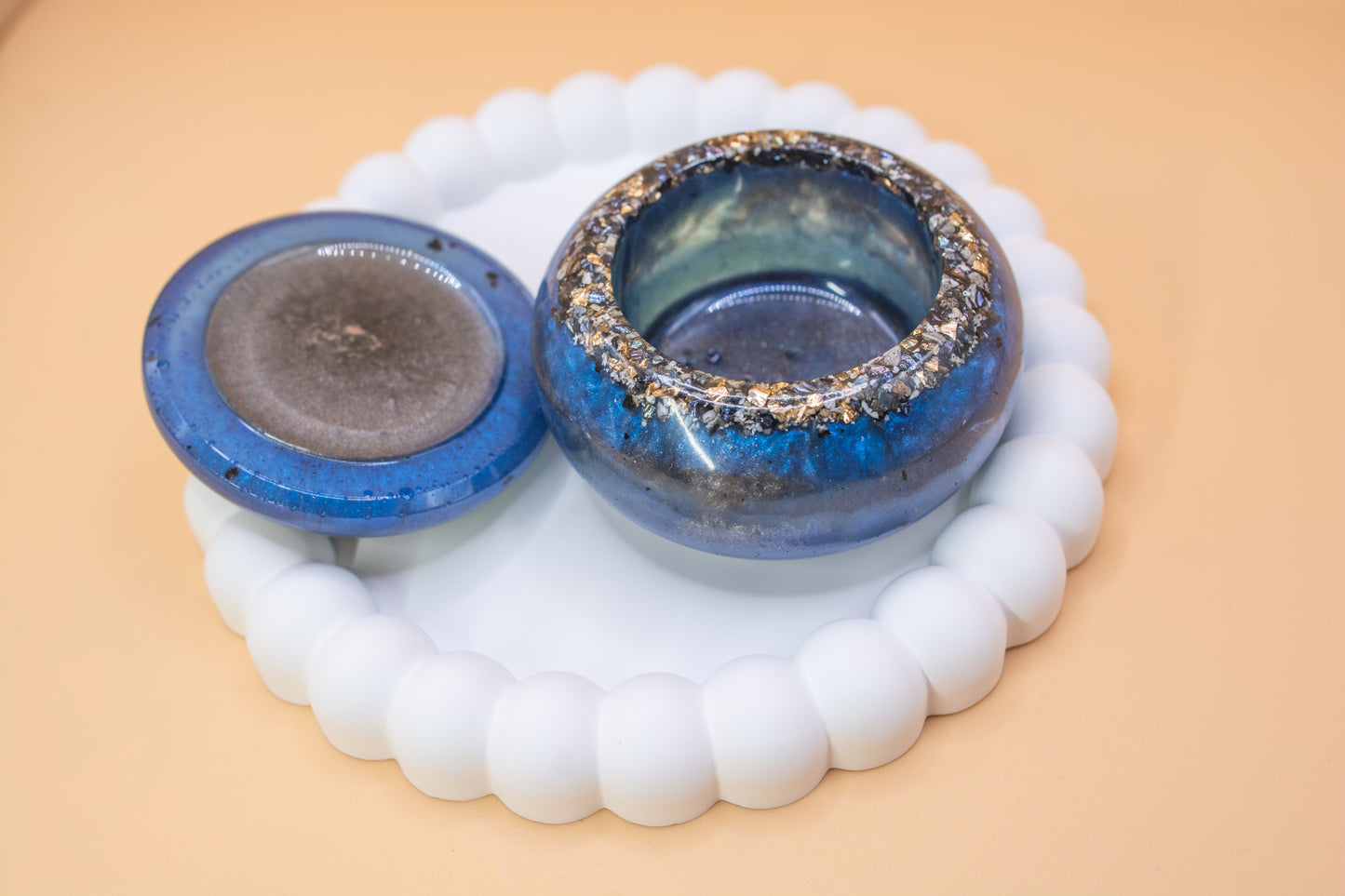 A front view of a metallic blue and grey handmade trinket pot, which has gold sparkle fragment and cremation ashes in it, the lid is to the side of the pot