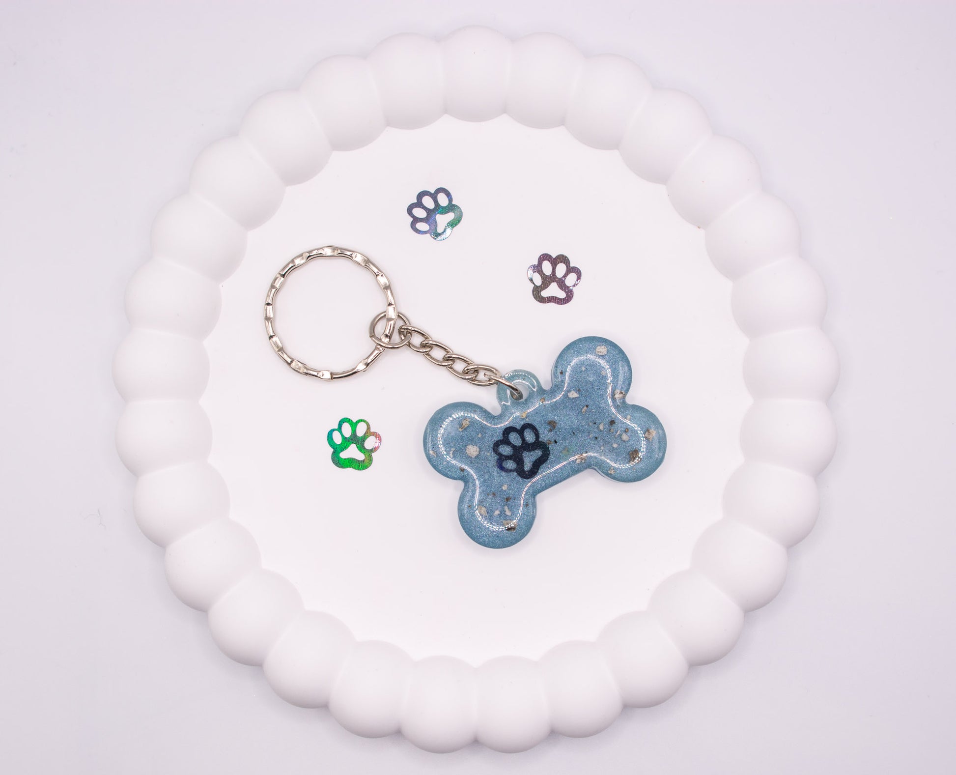 An aerial, overhead view of a light blue bone shaped keepsake memorial keyring, containing pet cremation ashes, set on a silver keychain