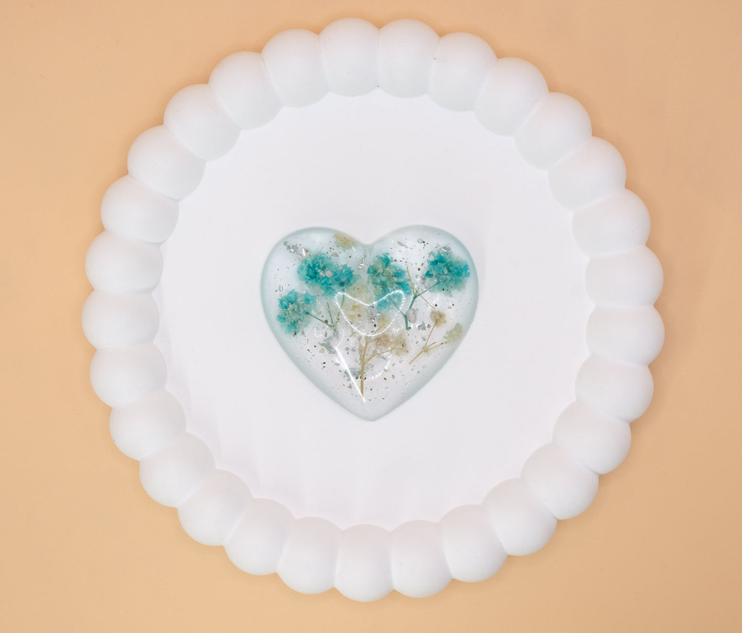 An overhead view of a heart shaped keepsake ornament, containing cremation ashes with blue and cream dried flowers and silver flakes, presented on a display tray