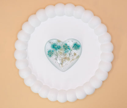An overhead view of a heart shaped keepsake ornament, containing cremation ashes with blue and cream dried flowers and silver flakes, presented on a display tray