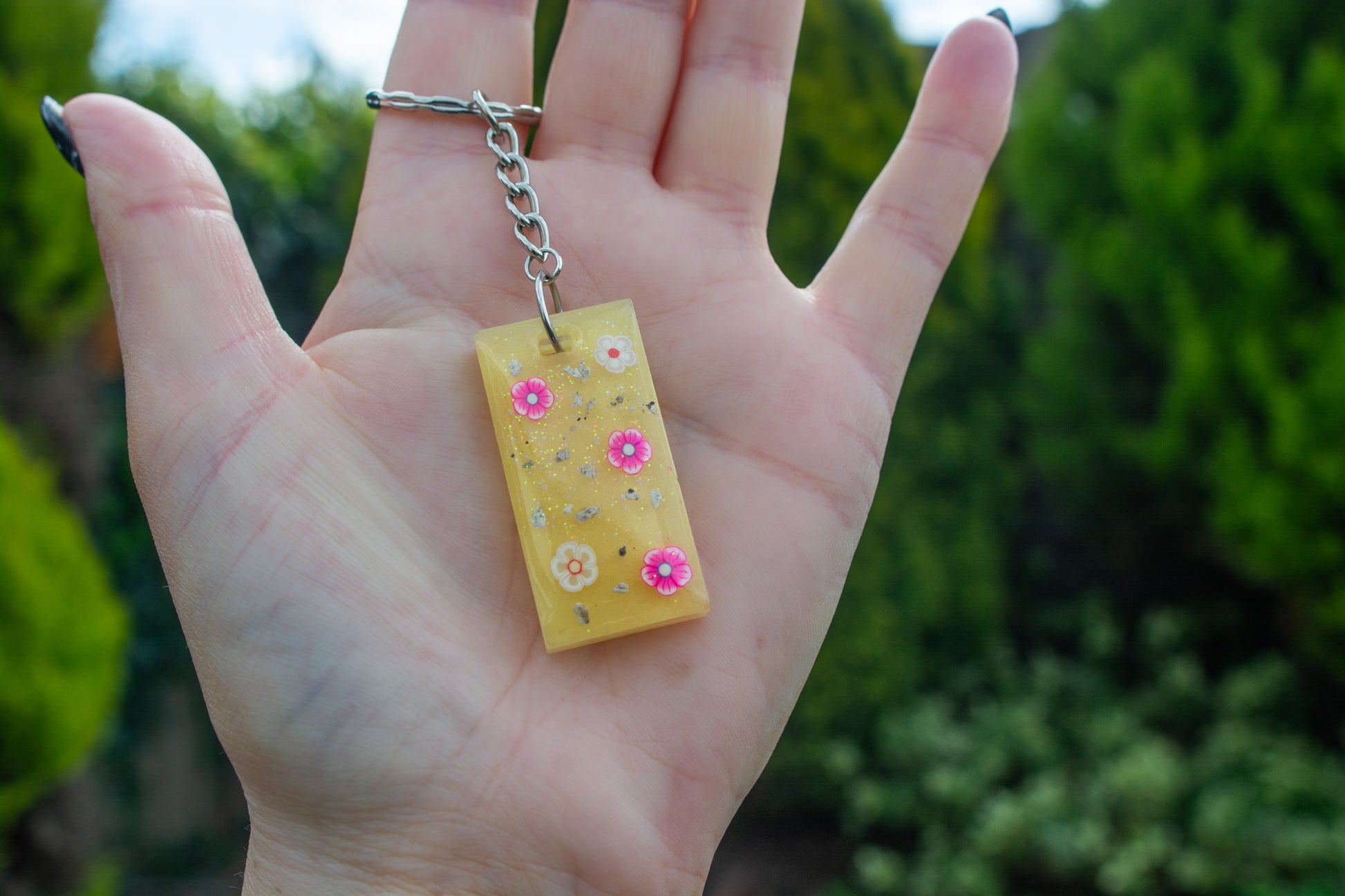 A hand holding a yellow rectangle shapedkeepsake memorial keyring outside, containing flower decorations and cremation ashes, set on a silver keychain