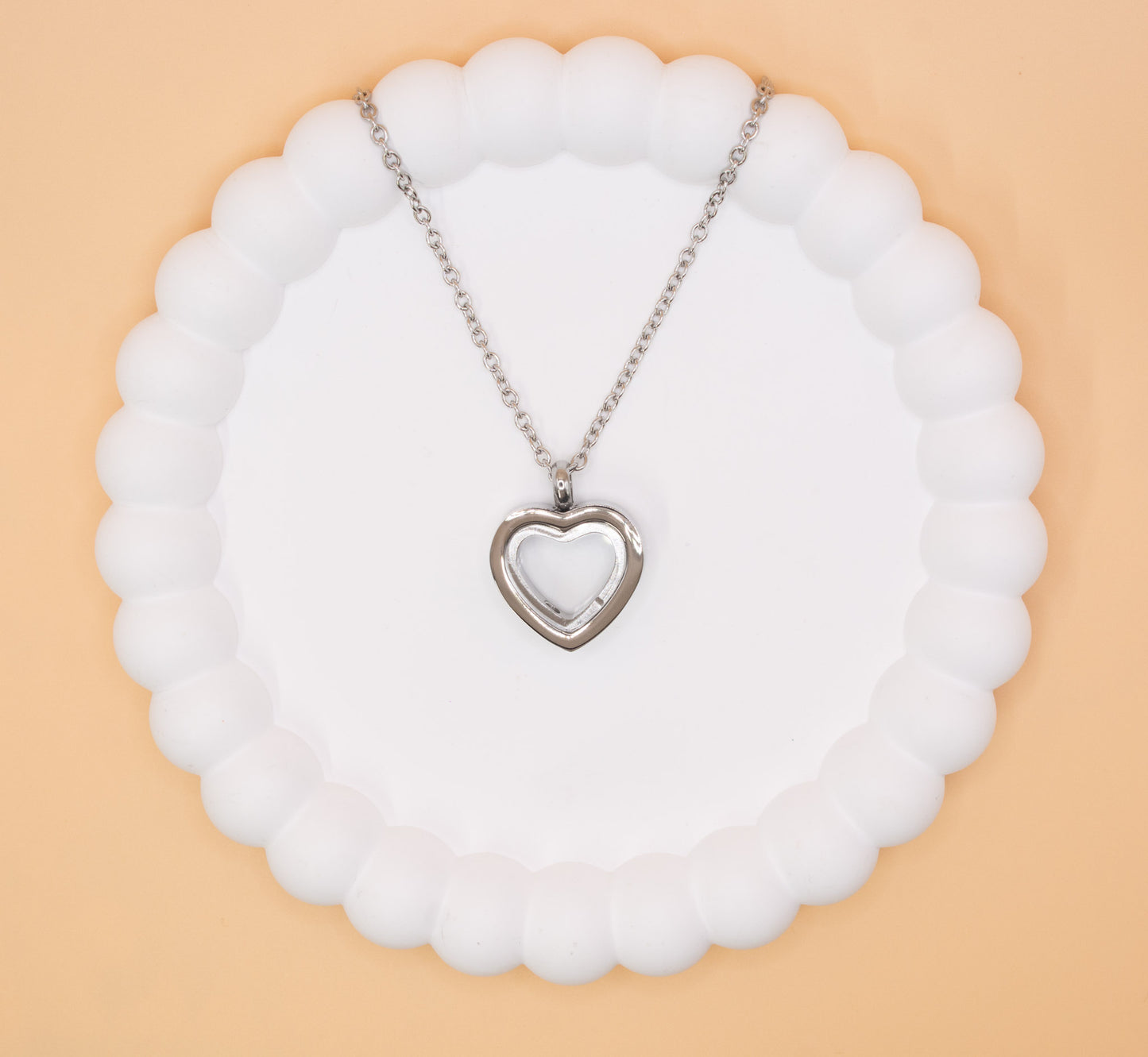 An aerial over head view of a stainless steel heart pendant, which can be self-filled with cremation ashes or sand from a favourite beach, it has a clear central window to see the contents
