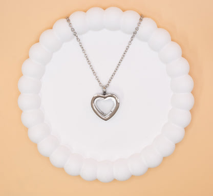An aerial over head view of a stainless steel heart pendant, which can be self-filled with cremation ashes or sand from a favourite beach, it has a clear central window to see the contents