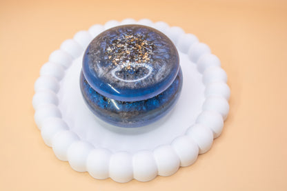 A front view of a metallic blue and grey handmade trinket pot, which has gold sparkle fragments and cremation ashes in it
