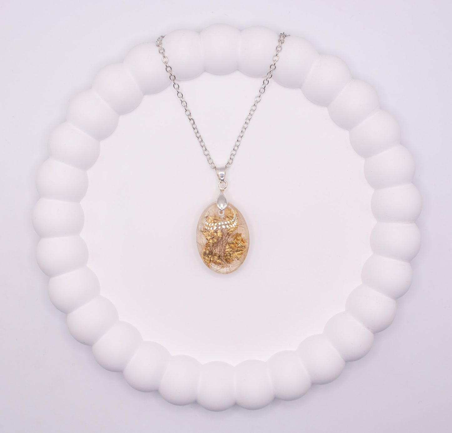 Over head view of a clear oval shaped keepsake memorial pendant, containing gold flake and human hair, set on a 925 sterling silver chain