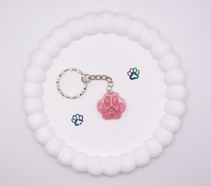 An over head view of a pink paw print shaped keepsake memorial keyring containing pet fur hair, set on a silver keychain