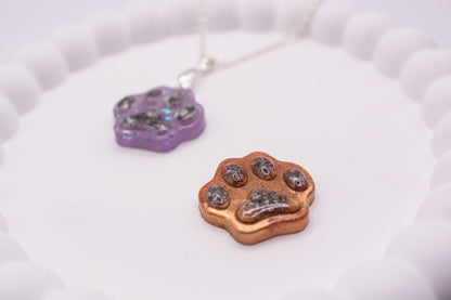 Close up angled view of One purple glitter and one metallic brown paw print keepsake memorial pendant, containing pet cremation ashes. The brown one is in focus and the purple one is out of focus.