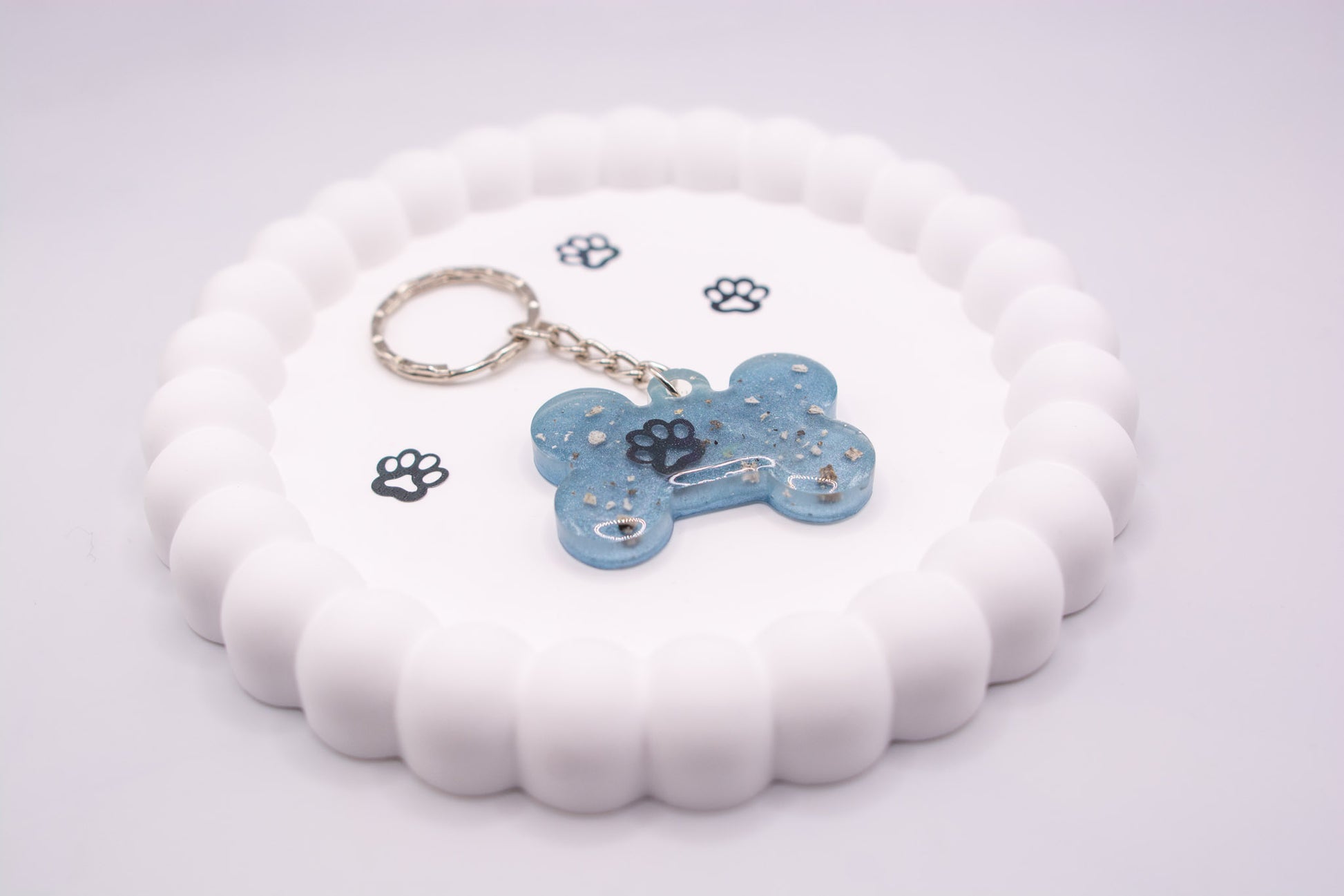 Angled view of a light blue bone shaped keepsake memorial keyring, containing a paw decoration and pet ashes, set on a silver keychain and presented on a display tray