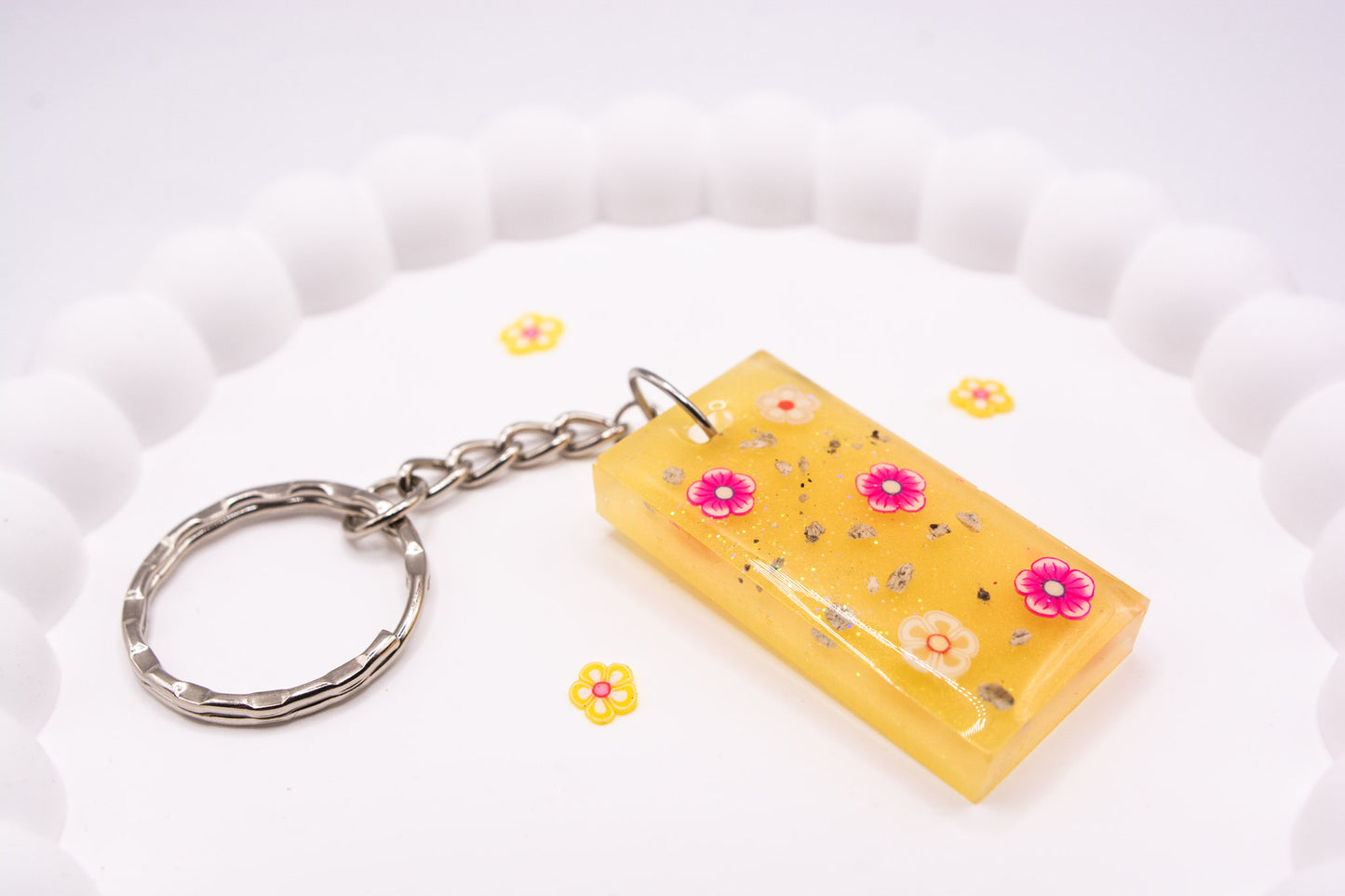 A close up angled view of a pretty yellow rectangle keepsake memorial keyring, containing flower decorations and cremation ashes, set on a silver keychain