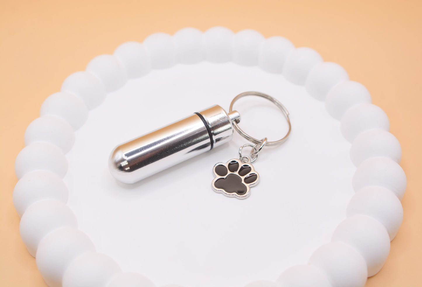 Displayed on a presentation tray is an angled close up view of a stainless steel silver keyring, with a black paw print charm. The keyring can be filled with a lost loved pet such as a cat or dog's cremation ashes